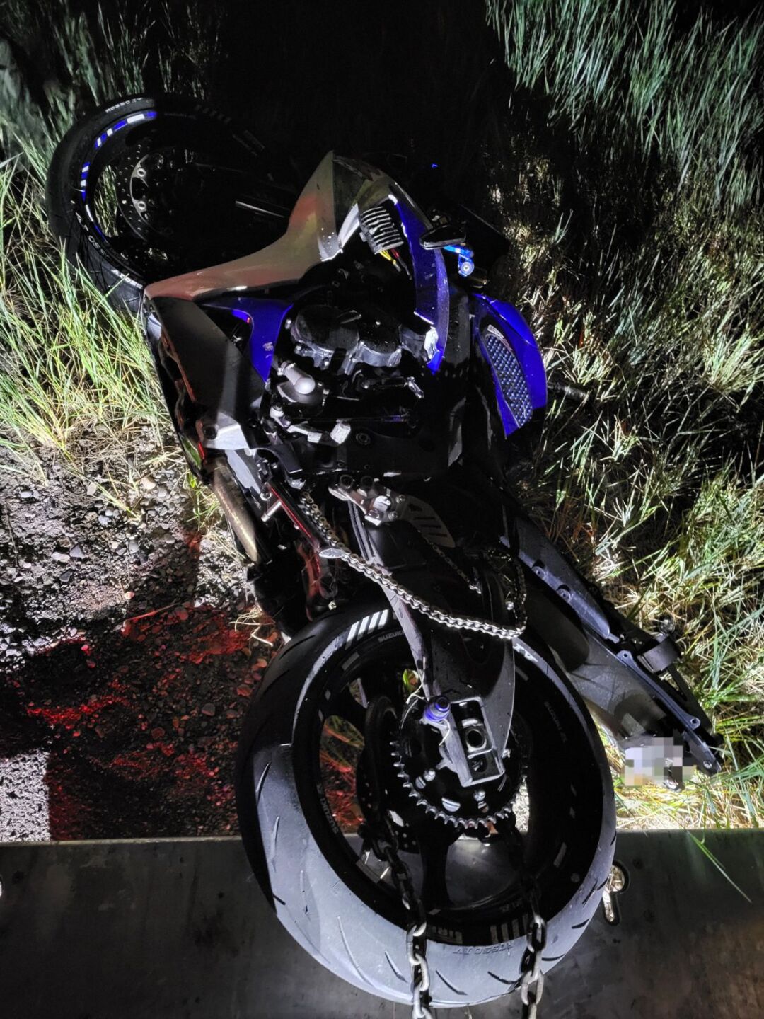 An image of the damaged blue 2007 Suzuki motorcycle involved in the crash released by Utah Highway Patrol. It's dark, so the image is light by the lights of a police cruiser from behind the photographer, resulting in shadows falling on the bike. It likes on its side in the grass next to the road.