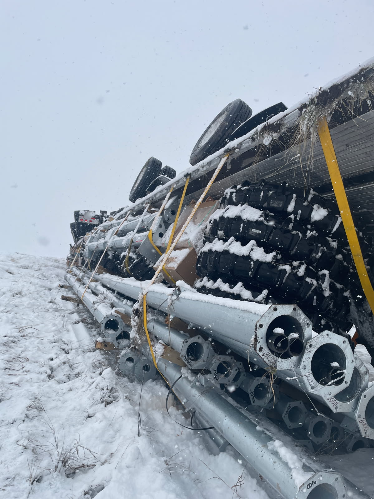 A photo from the scene released by Utah Highway Patrol. It's daytime, but snow covers the ground. The semi truck, hauling a load of pipes and tires is rolled over.