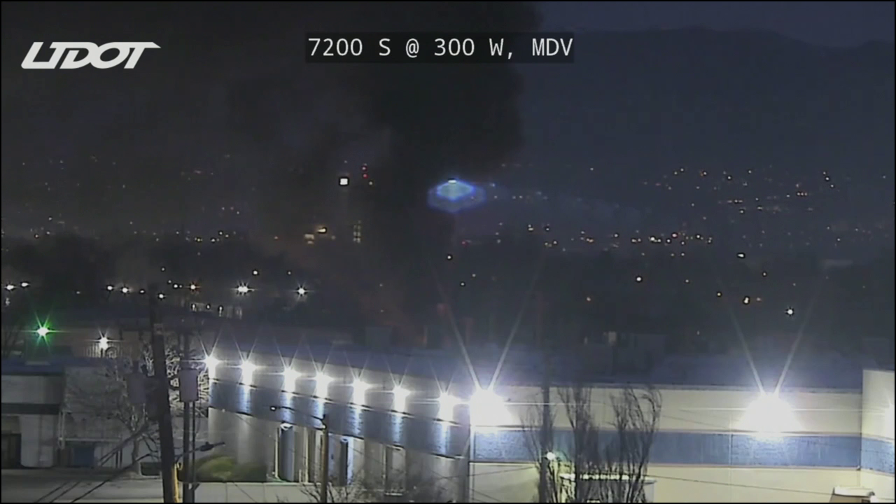 An image from a UDOT traffic camera. It is dark. Black smoke billows into the sky from behind a building as flood lights fill the image with light along the bottom of the frame.