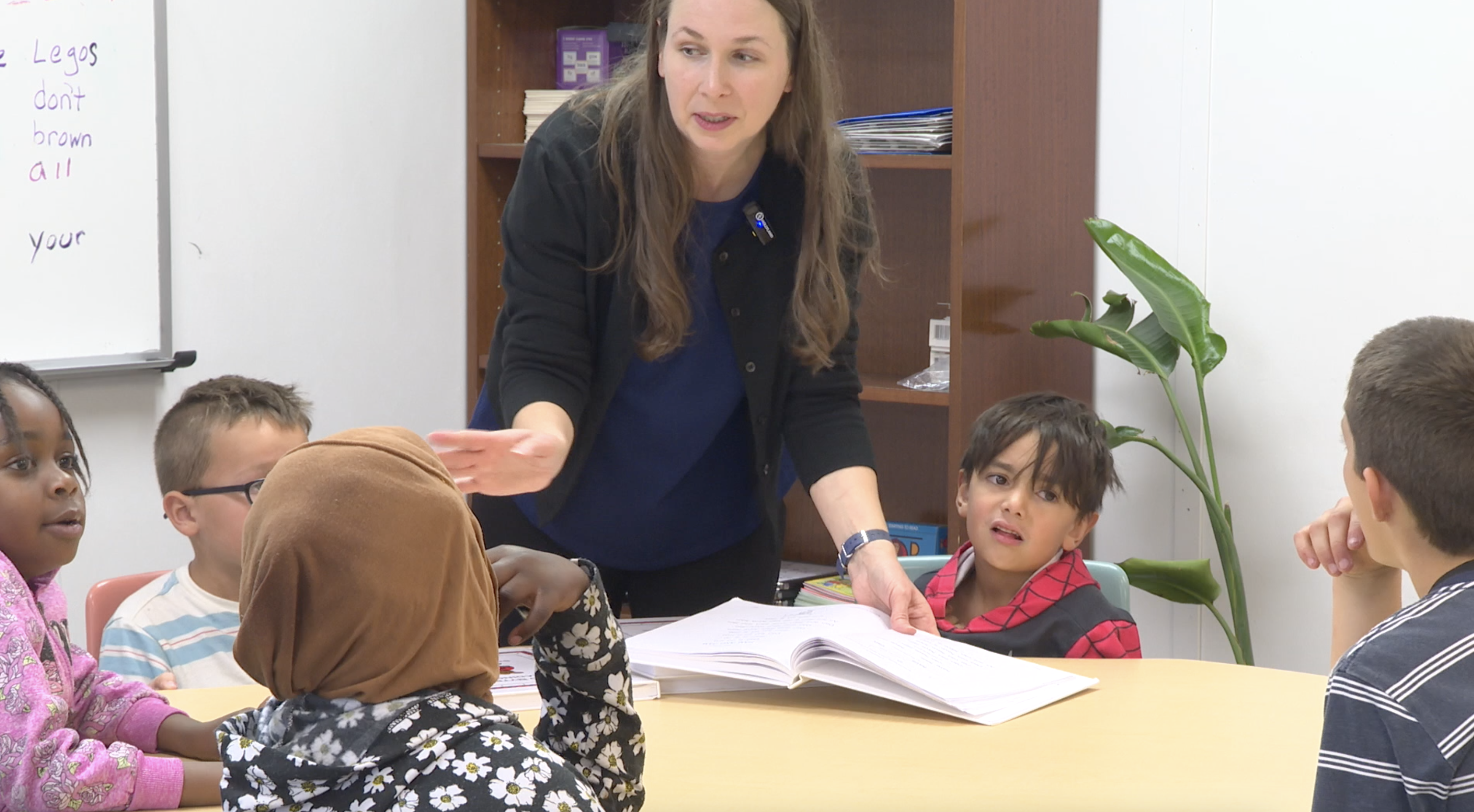 Students around a table with open books in front of them listen to a teacher give instructions
